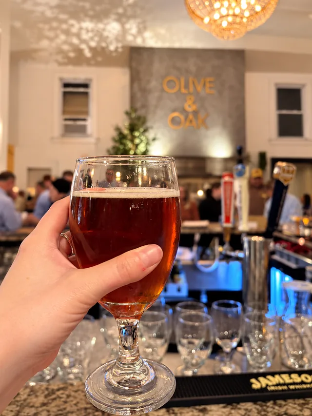 A glass of amber beer with the Olive & Oak bar and gold wall lettering behind it
